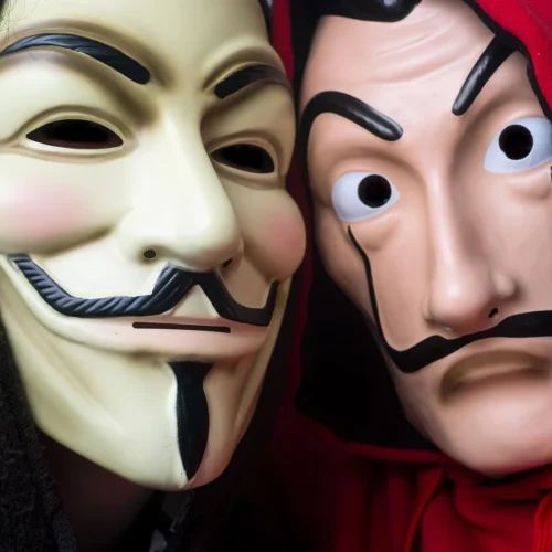Deux masques de visage posés l'un contre l'autre, l'un pâle à fine moustache et bouc, l'autre stylisé aux yeux exorbités sous une capuche rouge