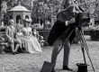 Une famille en costumes du XIXᵉ siècle pose dans un jardin devant un photographe penché sur un appareil photographique à soufflet