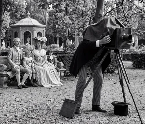 Une famille en costumes du XIXᵉ siècle pose dans un jardin devant un photographe penché sur un appareil photographique à soufflet