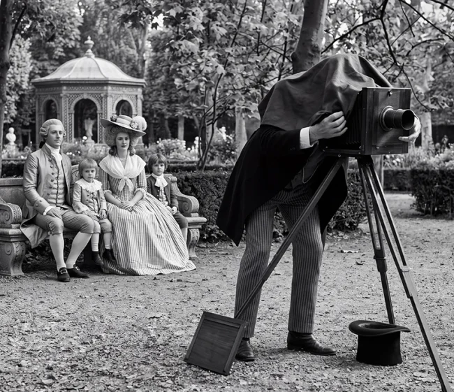 Une famille en costumes du XIXᵉ siècle pose dans un jardin devant un photographe penché sur un appareil photographique à soufflet