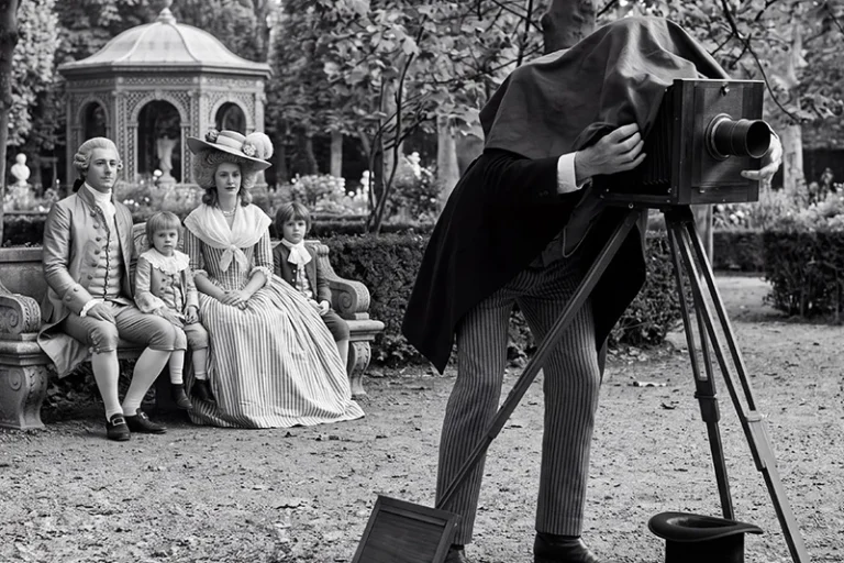 Une famille en costumes du XIXᵉ siècle pose dans un jardin devant un photographe penché sur un appareil photographique à soufflet