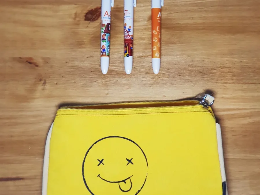 Trois stylos BIC 4 couleurs illustrés posés au-dessus d'une pochette jaune portant un smiley qui tire la langue et le logo AST67.