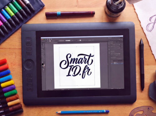Vue de dessus d'un bureau de graphiste : tablette graphique affichant le lettrage Smart ID, feutres Tria, stylo Rotring, encre de Chine, croquis au crayon et grattoir.