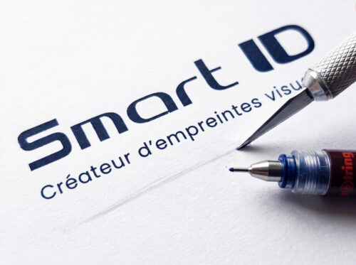 Logo Smart ID tracé à l'encre de Chine sur papier blanc, avec un stylo technique Rotring et la pointe d'un grattoir posés à côté.