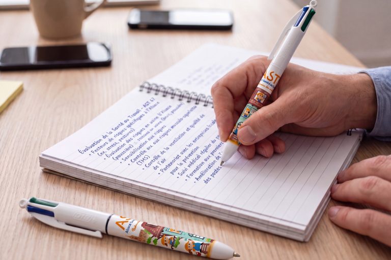 Main écrivant dans un carnet à spirale avec un stylo BIC 4 couleurs illustré aux couleurs d'AST67 ; un second stylo identique est posé sur le bureau.